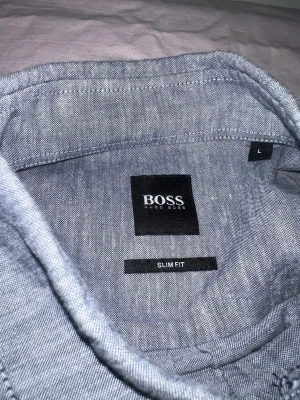 Grå slim fit skjorta från BOSS - Säljer en stilren grå skjorta från BOSS med slim fit passform. Skjortan har klassisk krage, knappar framtill och en diskret BOSS-logga på bröstet. Perfekt för dig som gillar en clean och modern look.