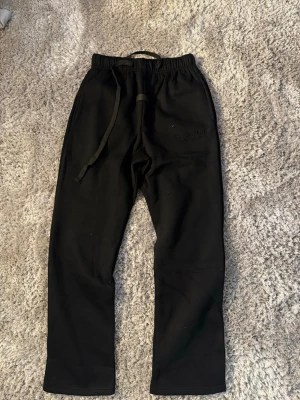 Svarta Essentials mjukisbyxor - Svarta mjukisbyxor från Fear of God Essentials med broderad logga på benet. Byxorna har resår i midjan och dragsko för perfekt passform. Tillverkade i mjukt material som är skönt att ha på sig. Perfekta för en avslappnad och trendig stil.