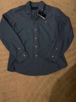 Blå skjorta från Ralph Lauren - Klassisk blå skjorta från Ralph Lauren med knappar framtill och button-down krage. Skjortan har lång ärm och den ikoniska rosa broderade loggan på bröstet. Tillverkad i mjuk bomull för en clean och tidlös look. Färgen ser lite ljusare ut än vad den är på bilden