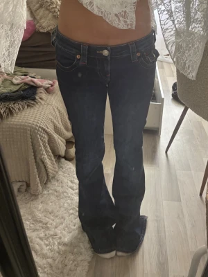Mörkblå lowwaist jeans True Religion - Säljer ett par mörkblå bootcut jeans från True Religion med kontrastsömmar och snygga detaljer på fickorna. Klassisk femficksmodell med låg midja och silverfärgade knappar. Perfekta för dig som gillar en tidlös och trendig look.