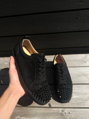 Louboutin Junior spikes  - Säljer ett par svarta sneakers i mockaimitation med coola svarta nitar på tån. Skorna har lågt skaft, svarta skosnören och en stilren design som sticker ut. Perfekta för dig som vill ha en edgy look till vardagsoutfiten. Size 40,5, bra skick med en liten defekt som syns på bild 6.