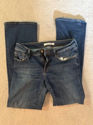 Blå raka jeans med slitningar - Säljer ett par klassiska blå jeans med raka ben och lätt slitna detaljer vid fickorna. Jeansen har fem fickor, bälteshällor och dragkedjegylf. Perfekta för en avslappnad och trendig look.