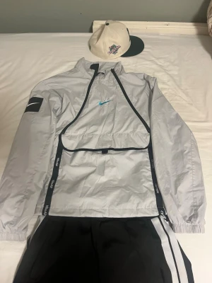 Nike grå vindjacka med svart detalj - Snygg grå vindjacka från Nike med svarta detaljer och blå Nike-logga på bröstet. Jackan har dragkedja, hög krage och stor ficka framtill. Elastiska muddar vid ärmslut och diskret Nike Air-tryck på ryggen. Perfekt för en sportig och trendig look.