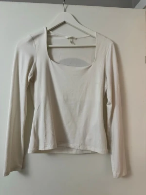Vit långärmad topp med fyrkantig ringning - En stilren vit långärmad topp med fyrkantig ringning. Toppen har en enkel och clean design, är figurnära och tillverkad i ett mjukt bomullsmaterial. Perfekt att matcha med jeans eller kjol för en fräsch look.