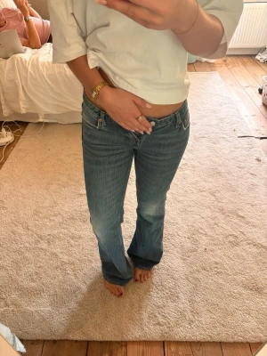 Blå bootcut jeans med låg midja - Säljer ett par blå bootcut jeans med låg midja och klassiska fickor både fram och bak. Jeansen har dragkedja och knapp framtill samt subtila slitningar för en cool vintagekänsla. Perfekta för dig som gillar retrostil och vill ha ett par riktigt snygga jeans.