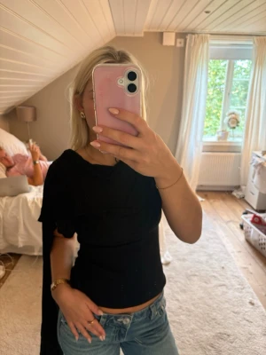 Svart asymmetrisk topp med knyt - Säljer en svart, tight topp med asymmetrisk design och ett långt knytband på ena axeln. Toppen är ärmlös på ena sidan och har kort ärm på andra, vilket ger en unik och trendig look. Perfekt att styla med jeans eller kjol.