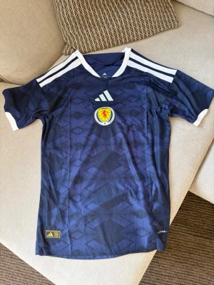 Skottland landslagströja – Adidas - Mörkblå Skottland fotbollströja från Adidas med klassiskt geometriskt mönster, vita Adidas-ränder över axlarna och Skottlands landslagsmärke framtill. Slim fit-modell med AEROREADY/HEAT.RDY-liknande funktionsmaterial och Authentic Licensed Product-märkning nedtill.  Fint skick utan tydliga större skador eller fläckar. Storlek syns inte tydligt på bilderna.