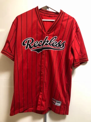Röd randig skjorta Reckless - Säljer en röd skjorta med svarta vertikala ränder och texten 'Reckless' broderad över bröstet. Skjortan har korta ärmar, knappar framtill och sportig basebollstil. Perfekt för dig som vill sticka ut med en streetwear-look.