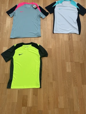  3 pack - Nike sporttröjor i olika färger ”Äkta” - Tre sporttröjor från Nike i olika färger: en ljusgrå med rosa och svarta detaljer, en vit med turkosa och svarta detaljer samt en neongul med svarta detaljer. Alla har korta ärmar och klassisk sportig design, perfekta för träning eller match.