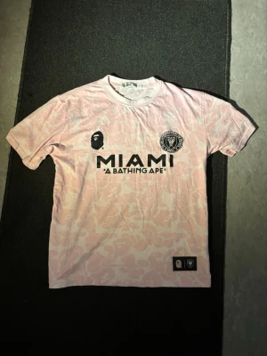 BAPE t-shirt - Säljer en limited edition rosa MIAMI t-shirt från A Bathing Ape med svart logga och tryck framtill. T-shirten har korta ärmar, rund hals och ett subtilt kamouflagemönster i ljusrosa toner. Perfekt för dig som gillar streetwear och unika samarbeten. Storlek L men passar mer som M/S