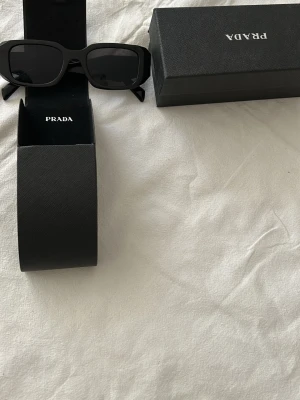 Svarta solglasögon från Prada - Stilrena svarta solglasögon från Prada med rektangulära bågar. Klassisk design med breda skalmar och diskret logga på sidan. Kommer med originaletui och låda. Perfekta för dig som vill ha en tidlös accessoar med lyxig känsla.
