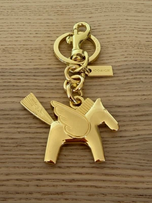 Guldfärgad Pegasus nyckelring Coachh - Säljer en guldfärgad nyckelring från Coachh i form av en Pegasus med vingar och svans. Tillverkad i metall med blanka detaljer och Coachh-logga på brickan. Perfekt att hänga på väskan eller nycklarna för en unik touch.