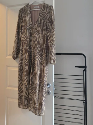 Zebramönstrad långklänning Adoore - Snygg långklänning med zebramönster i beige och grå toner. Klänningen har v-ringning, långa ärmar och knappar framtill. Perfekt för dig som vill sticka ut med ett djurmönster. Tillverkad i ett lätt och luftigt material.
