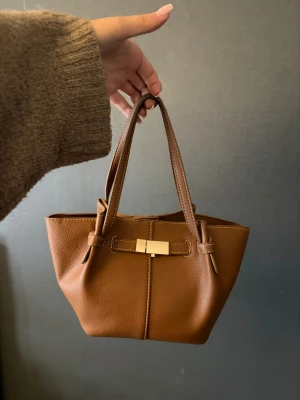 Brun handväska med gulddetalj - Snygg brun handväska i konstläder med två handtag och guldfärgad detalj framtill. Väskan har en stilren, lätt fyrkantig form och praktiskt innerfack med dragkedja. Perfekt storlek för det viktigaste och enkel att matcha till olika outfits. Man får även med ett långt band till väskan 