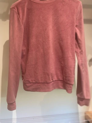 Dammig rosa sweatshirt med struktur - En stilren sweatshirt i dammigt rosa med diskret strukturmönster över hela tröjan. Tröjan har rund halsringning, långa ärmar och ribbade muddar vid ärmslut och nederkant. Perfekt för en avslappnad och trendig look.