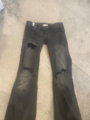 Svarta  jeans från Gina Tricot - Säljer ett par svarta jeans från Gina Tricot med slitningar och hål på båda benen. Jeansen har raka ben, klassisk femficksmodell och knappgylf. Perfekta för en avslappnad och trendig look.