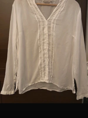 Vit blus med volangdetaljer - Säljer en vit blus med v-ringning och dekorativa volangdetaljer längs knappraden framtill. Blusen har långa ärmar och lätt avslappnad passform. Perfekt för en stilren och enkel look.