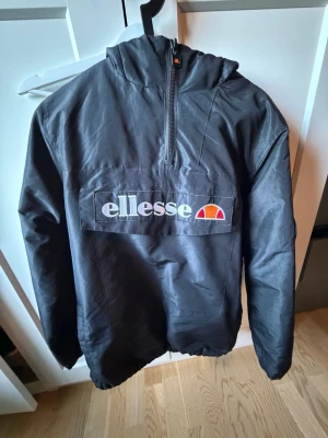 Svart vindjacka från Ellesse - Snygg svart vindjacka från Ellesse med halv dragkedja och stor logga på bröstet. Jackan har huva och elastisk nederkant, perfekt för blåsiga dagar. Materialet är lätt och vindtätt, vilket gör den enkel att bära och matcha till olika outfits.