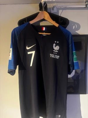 Frankrike fotbollströja Griezmann #7 - Säljer en officiell blå fotbollströja från Nike med Griezmann och nummer 7 på ryggen. Tröjan har vita detaljer, två stjärnor och FFF-logga på bröstet samt VM 2018-detaljer på ärmen. Perfekt för dig som älskar landslagsfotboll och vill stötta Frankrike.
