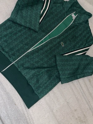 Grön mönstrad hoodie från Lacoste - Snygg grön hoodie från Lacoste med diskret geometriskt mönster och dragkedja framtill. Har vita och mörkgröna ränder på ärmarna och den klassiska krokodilloggan på bröstet. Tillverkad i mjukt material som känns skönt mot huden. Perfekt för en chill och sportig look.