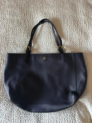 Svart läderväska med gulddetaljer från Tory Burch - Stilren svart handväska i slätt läder med justerbara axelband och små gulddetaljer. Väskan har en enkel, rymlig design och ett diskret metallogo framtill. Perfekt för dig som gillar klassisk och tidlös stil.