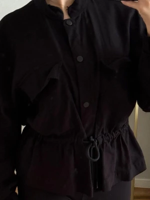 Svart overshirt med snörning - Snygg och mångsidig svart jacka/överskjorta från Zara. Den har dragsko i midjan så att man kan anpassa passformen, tryckknappar framtill, stående krage och två rymliga bröstfickor. Ärmarna har elastiska slut. Passar perfekt som en lätt jacka på våren/sommaren eller som överskjorta inomhus under svalare dagar!