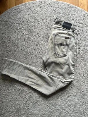 Grå nudie jeans, pipe led - Snygga grå jeans med slim passform och svarta detaljer vid bakfickan. Klassisk femficksmodell med diskreta sömmar och stilren look. Perfekta för dig som gillar en enkel men trendig stil.