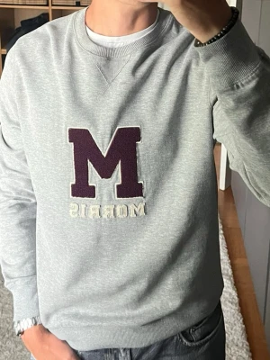 Morris sweatshirt med M-märke i grå - stl M - Morris sweatshirt med M-märke i gråmelerad, storlek M. 80% bomull, 20% polyester, ruggad och mjuk på insidan. Stort M och Morris-text påsytt fram. Personen på bilden är 189cm, väger 78kg.  Använd men i fint skick, ett hål som har reparerats, visas på bild 6.  Rök- och djurfritt hem. Skriv gärna om du vill ha fler bilder eller undrar något!