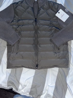 Grå cardigan från Moncler - Snygg grå dunväst från Moncler med ribbstickade detaljer på krage och ärmhål. Västen har dragkedja framtill, två sidofickor och Moncler-logga på axeln. Perfekt för lager-på-lager och ger en clean, modern look.