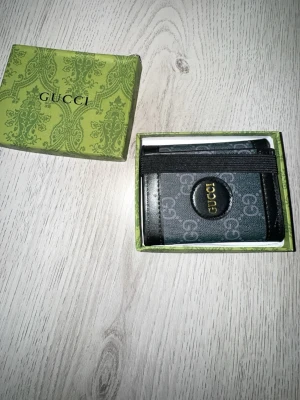 Svart  plånbok med monogram - Snygg svart plånbok från  med klassiskt monogram i grått och en rund logga framtill i guld. Plånboken har elastiskt band och levereras i original grön box med mönster. Perfekt för kort och sedlar.