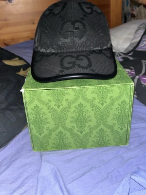 Svart keps från Gucci med GG-mönster - Säljer en svart keps från Gucci med det klassiska GG-mönstret i mörkgrått över hela kepsen. Kepsen har böjd skärm och justerbart band bak. Materialet är canvas med detaljer i läder på skärmen. Perfekt för dig som vill ha en stilren och lyxig accessoar.