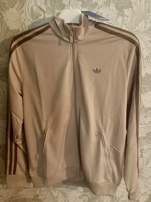 Beige Adidas track jacket med bruna ränder - Snygg beige track jacket från Adidas originals med bruna ränder längs ärmarna och Adidas logga på bröstet. Dragkedja framtill och två fickor med dragkedja. Tillverkad i ett mjukt och stretchigt material som passar perfekt till träning eller chill. Helt ny med prislapp. I storlek 164cm.