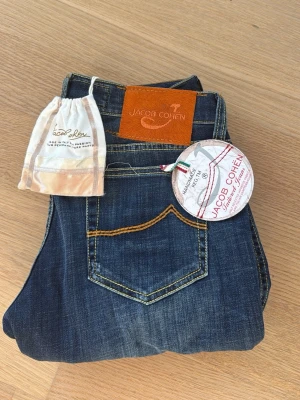 Mörkblå jeans från Jacob Cohen - Snygga mörkblå jeans från Jacob Cohen med raka ben och kontrastsömmar i gult. Klassisk femficksmodell med patch i brunt läder bak. Tillverkade i mjukt denimtyg, perfekta för en avslappnad men stilren look. Helt nya tags kvar! Pris kan diskuteras!!!