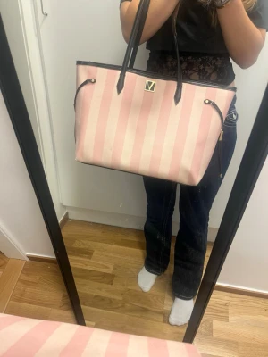Victoria Secret Rosa randig tote handväska - Snygg rosa och ljusrosa randig tote handväska med svarta handtag och detaljer. Väskan har en rektangulär form och ett metallmärke framtill. Perfekt storlek för att få plats med allt du behöver. Materialet ser ut att vara slitstark textil.