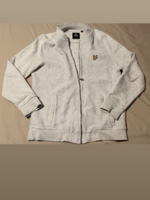 Ljusgrå ziptröja från Lyle & Scott - Ljusgrå ziptröja från Lyle & Scott med klassisk gul logga på bröstet. Tröjan har ståkrage, ribbade muddar och två fickor framtill. Materialet är mjukt och strukturerat, perfekt för en avslappnad stil.