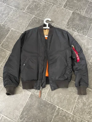 Svart bomberjacka Alpha Industries stl M - Klassisk svart bomberjacka från Alpha Industries med orange innerfoder och ribbade muddar vid ärmslut och midja. Jackan har två snedställda fickor med tryckknappar och en ikonisk röd tag på ärmfickan. Tillverkad i slitstarkt nylonmaterial. 