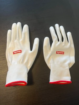 Vita handskar från Supreme - Säljer ett par vita handskar från Supreme med röd logga på ovansidan och röd kant vid handleden. Handskarna är tunna och smidiga, perfekta för dig som vill ha en clean och stilren accessoar.