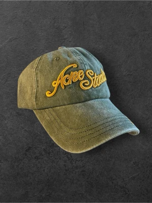 Olivgrön Acne Studios keps - Snygg olivgrön keps från Acne Studios med gul broderad logga framtill och på skärmen. Klassisk böjd skärm och justerbar rem bak. Tillverkad i slitstark bomull som ger en avslappnad och trendig look.