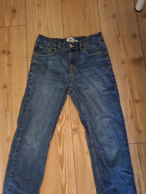 Blå raka jeans med fem fickor - Klassiska blå jeans i rak modell med fem fickor och dragkedja gylf. Jeansen har en lätt tvättad look och är tillverkade i slitstarkt denimtyg. Perfekta för en avslappnad och stilren vardagsoutfit.