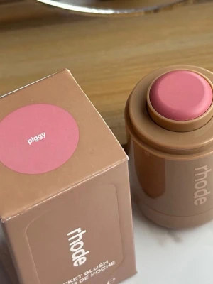Rhode Pocket Blush Piggy - Krämrouge från Rhode i färgen Piggy, en mjuk rosa nyans. Kommer i en rund, beige förpackning med tryckknapp och matchande kartong. Perfekt för att ge kinderna en naturlig, fräsch glow och enkel att ta med i väskan.