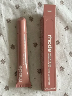 rhode Peptide Lip Tint Toast - Säljer ett rhode Peptide Lip Tint i färgen Toast. Läppglanset kommer i en mjuk tub med skruvkork och har en varm, nude-brun ton. Förpackningen är stilren och modern med vit text. Innehåller 10 ml.