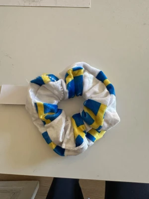 Scrunchie med svensk flagga-mönster - En scrunchie i glansigt tyg med mönster inspirerat av svenska flaggan. Färgerna är blå, gul och vit, och materialet känns mjukt och elastiskt. Perfekt accessoar för att piffa upp håret med lite svensk känsla.