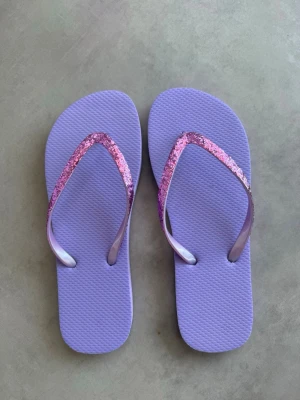 Lila flip-flops med glitterremmar - Säljer ett par lila flip-flops med glittriga remmar. De är i mjukt, flexibelt gummimaterial som är perfekt för sommaren. Snygga att matcha med shorts, kjol, jeans eller klänning för en färgglad coolare look. Dem är helt nya/oanvända!