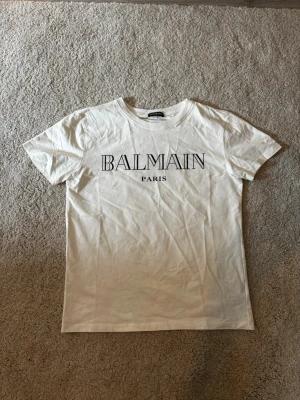 Vit t-shirt från Balmain Paris - Snygg vit t-shirt från Balmain Paris med klassisk rund hals och tryckt logga framtill. Tillverkad i mjukt bomullsmaterial och har en avslappnad passform. Perfekt för dig som gillar stilrena och trendiga plagg.