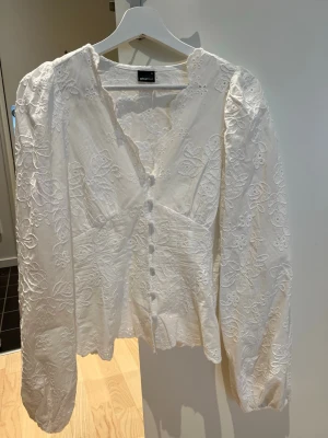 Vit broderad blus från Gina Tricot - Supersöt vit blus från Gina Tricot med broderade blommor, v-ringning och klädda knappar framtill. Blusen har puffiga långärmar och lätt utsvängd nederkant. Perfekt för dig som gillar romantisk och trendig stil.