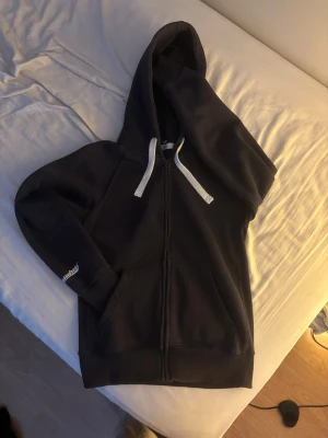 Navy blue russedress hoodie  - Navy blå hoodie från Russemerch med dragkedja och vita snören i huvan. Klassisk modell med kängurufickor framtill och ribbade muddar. Tryckt Russedress-logga på ena ärmen.