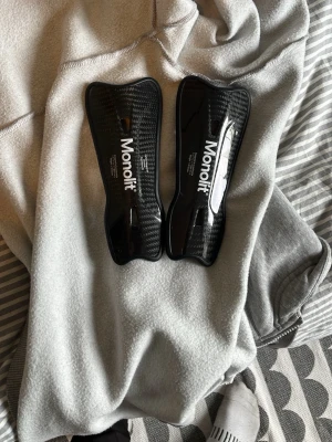 Svarta benskydd från Monolit - Säljer ett par svarta benskydd från Monolit med kolfibermönster. De är lätta och smidiga, perfekta för fotboll eller andra sporter där skydd behövs. Snygg design med vit logga på framsidan.
