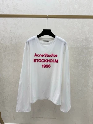 Vit långärmad topp Acne Studios - Vit långärmad topp från Acne Studios med trycket 'Acne Studios STOCKHOLM 1996' i rosa framtill. Klassisk rund halsringning och avslappnad passform. Perfekt för dig som gillar stilrena plagg med en twist.