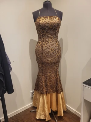 Guldig långklänning med spetsdetaljer - Elegant långklänning i guld med smala axelband och figurnära passform. Klänningen har ett lager av svart spets med blommigt mönster över ett glansigt guldtyg och en utsvängd nederdel i satinliknande material. Perfekt för speciella tillfällen. Stl 36-38, bröst ca 76-80, midja ca 74cm, höft ca 100cm, längd ca 130cm från armhåla.