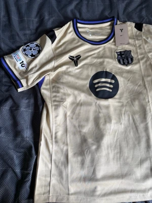 Gul FC Barcelona matchtröja Nike - Snygg gul FC Barcelona matchtröja från Nike med svarta detaljer och tryck. Tröjan har klubbmärke, Spotify-logga och Champions League-märke på ärmen. Tillverkad i lätt och ventilerande Dri-FIT-material, perfekt för fotboll eller träning.